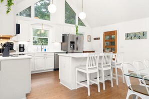 A modern kitchen with white cabinetry, a central island with seating, and a dining area with white chairs. - Waters Edge| Boat Lift+Fenced Yard+Kayaks+PingPong (Littleton)