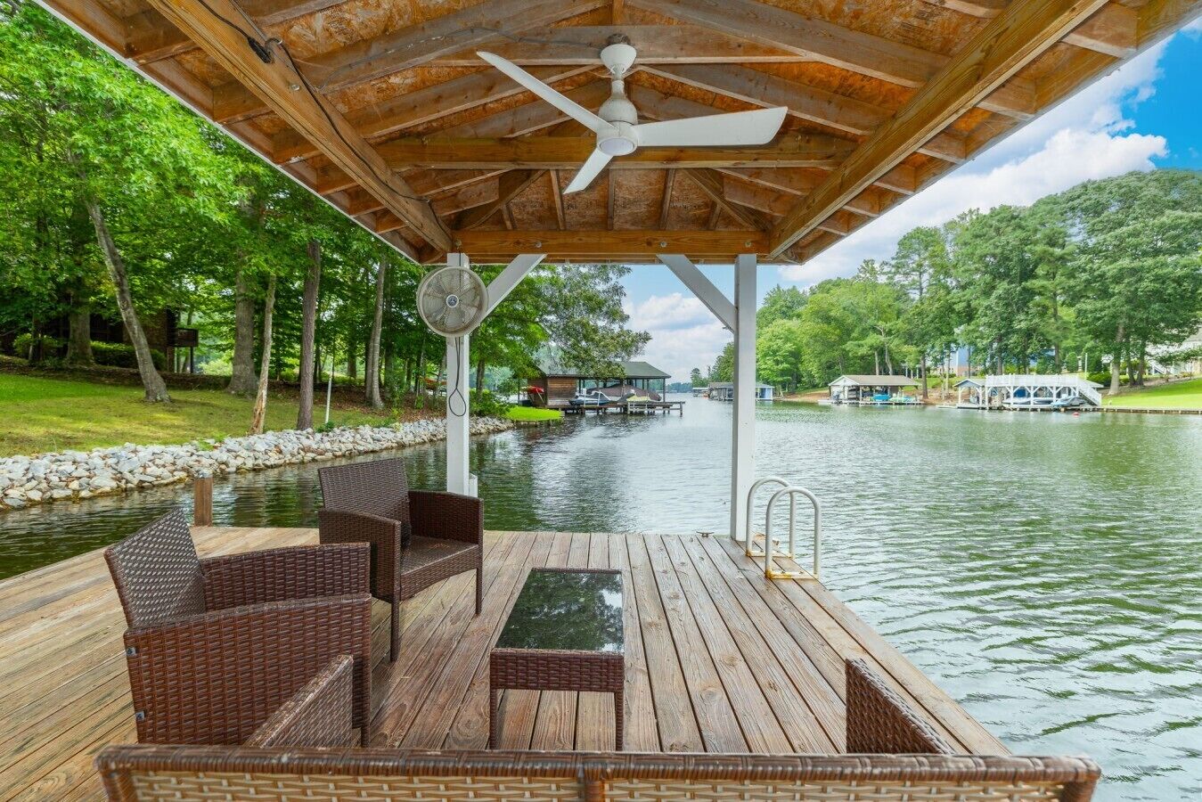A covered deck with wicker furniture, a ceiling fan, and a view of a lake with docks and boats.