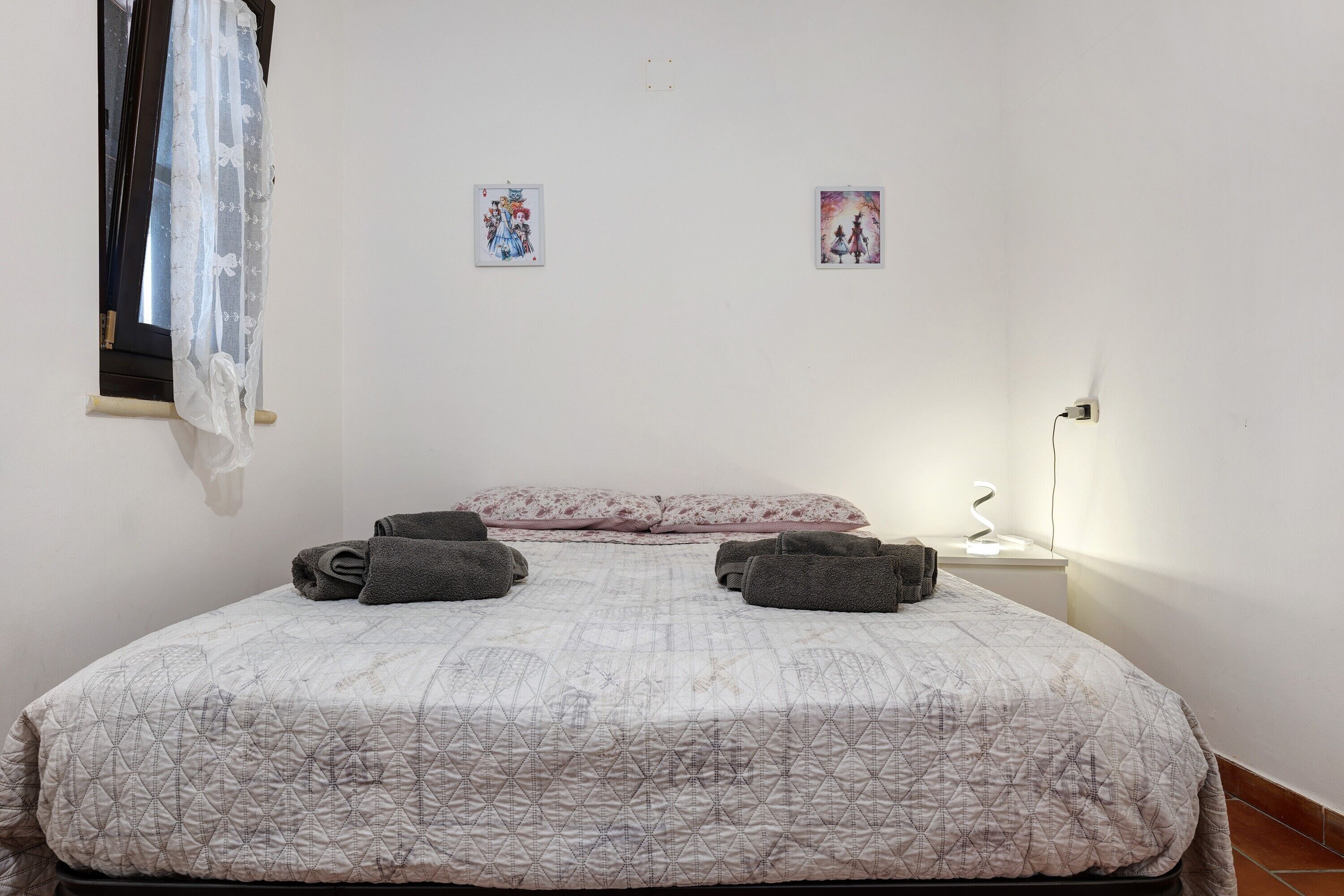Un dormitorio con una cama, dos almohadas, una ventana con cortina y dos cuadros en la pared.