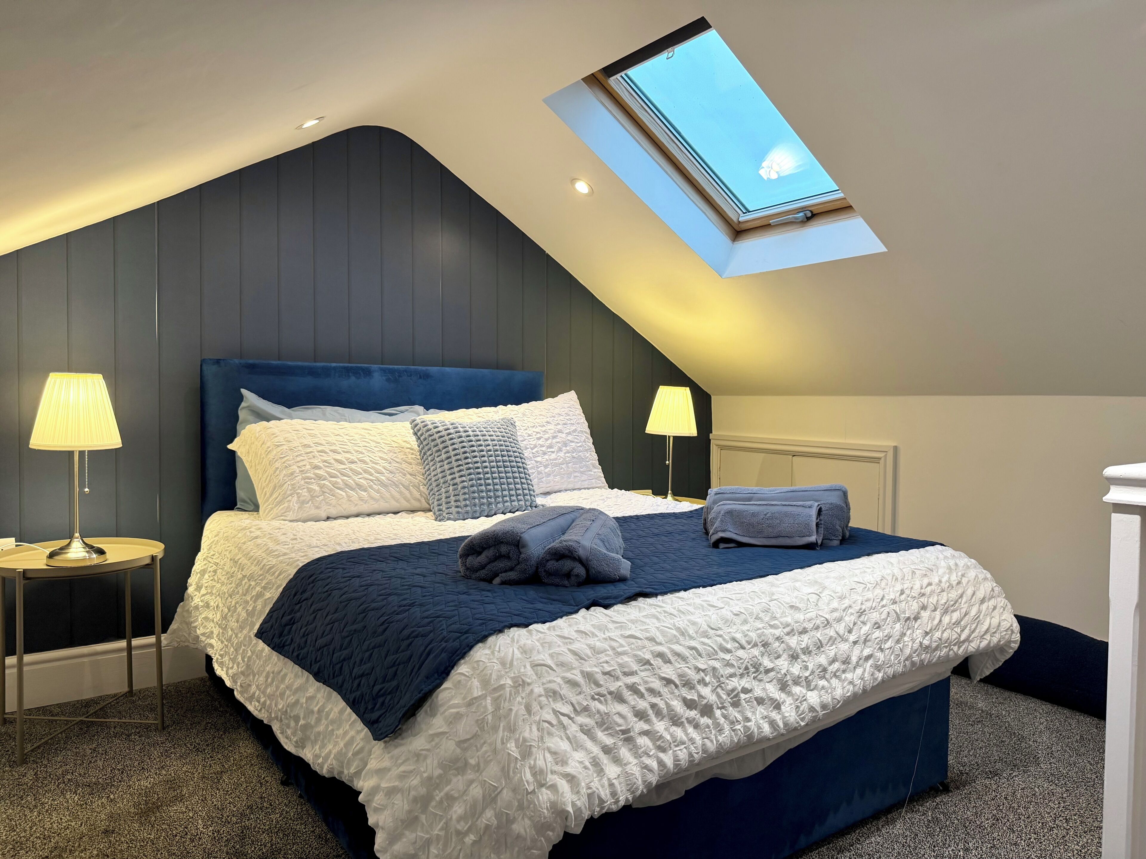 A bedroom with a sloped ceiling, a skylight, a bed with a blue headboard, two bedside lamps, and a neatly made bed with a blue and white quilt.
