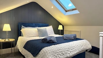A bedroom with a sloped ceiling, a skylight, a bed with a blue headboard, two bedside lamps, and a neatly made bed with a blue and white quilt.