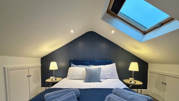 Ein Schlafzimmer mit schrägen Wänden, einem Oberlicht, einem großen Bett mit blauer Kopfteil, zwei Nachttischen mit Lampen sowie einem Kleiderschrank.