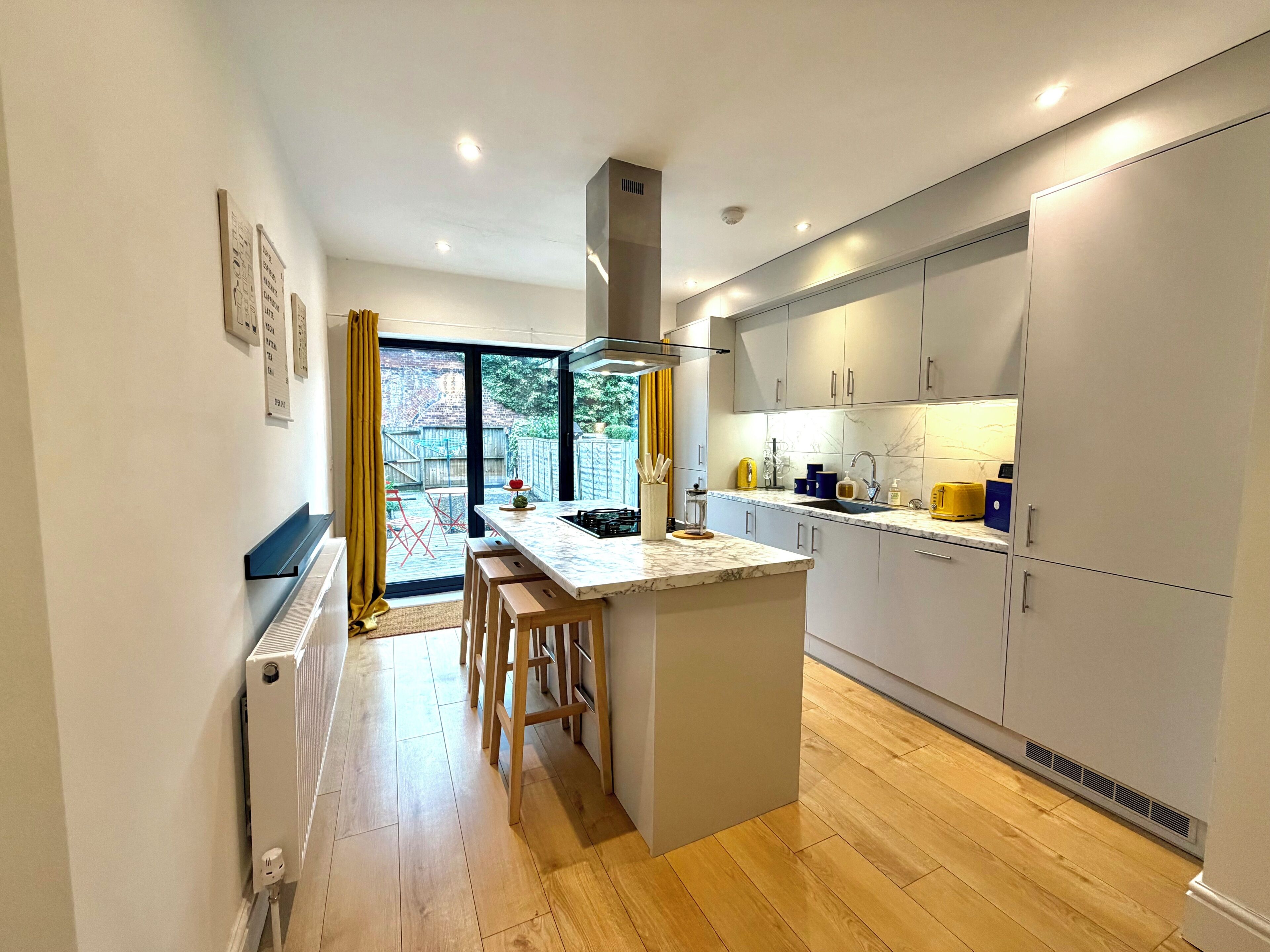 A modern kitchen with a central island, wooden stools, and a view of the backyard through large windows.