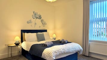 Ein Schlafzimmer mit einem Bett, zwei Nachttischlampen, einem Wandbild und einem Fenster mit Jalousien.