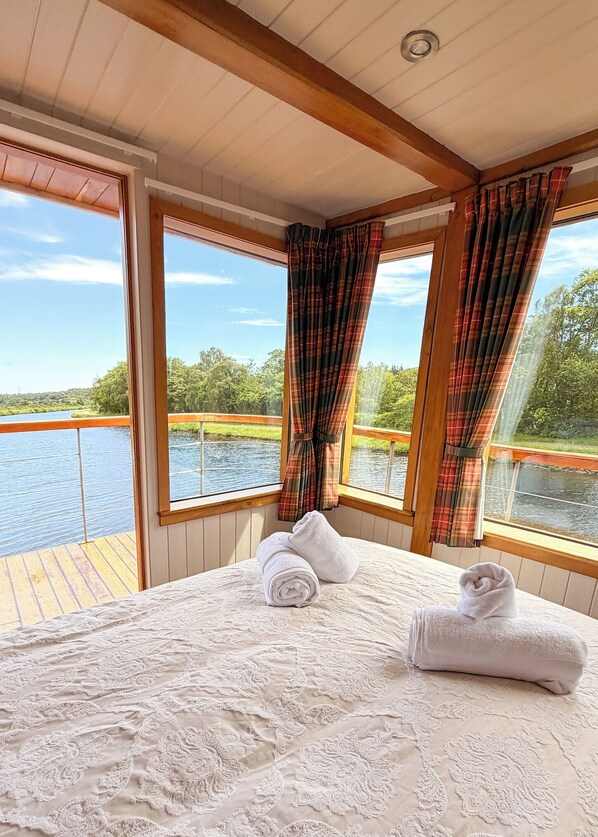 A bedroom with a large bed, two windows with curtains, a view of a river, and a wooden deck outside.