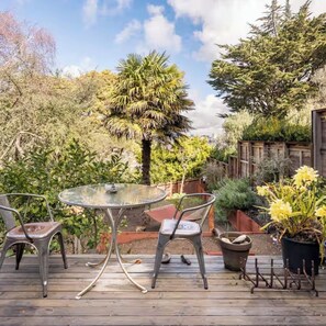 A wooden deck with a glass table and metal chairs, surrounded by various plants and trees.