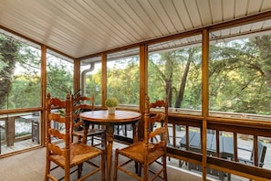 A conservatory with a round table and four chairs, large windows, and a view of a wooded area.