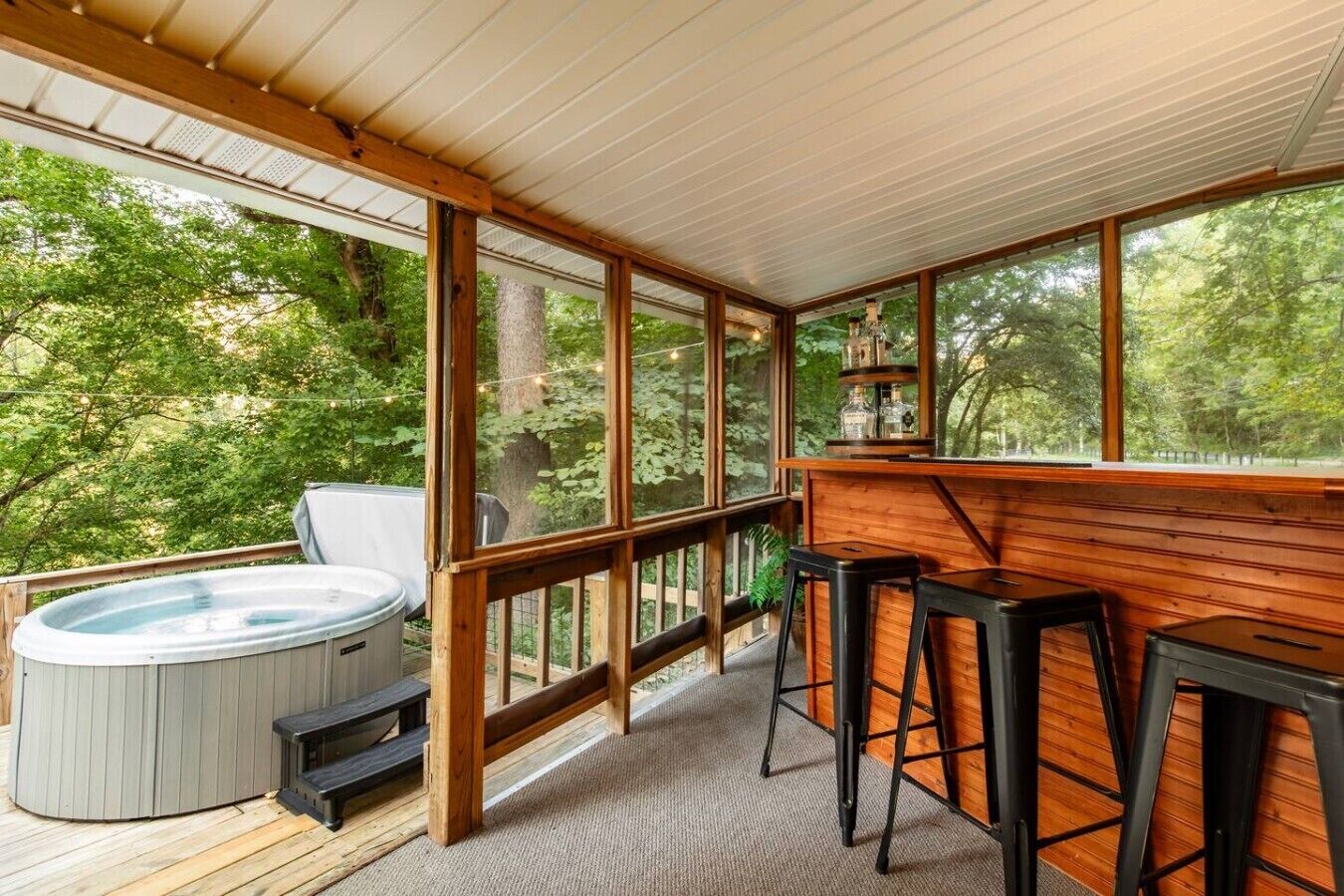 A wooden deck with a hot tub, bar stools, and a view of a forest.