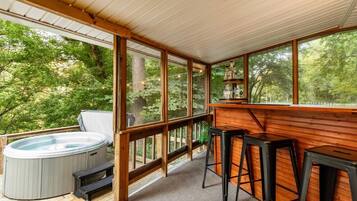 A wooden deck with a hot tub, bar stools, and a view of a forest.