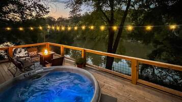 Un bain à remous sur une terrasse en bois, agrémentée de guirlandes lumineuses, avec vue sur une étendue d’eau.