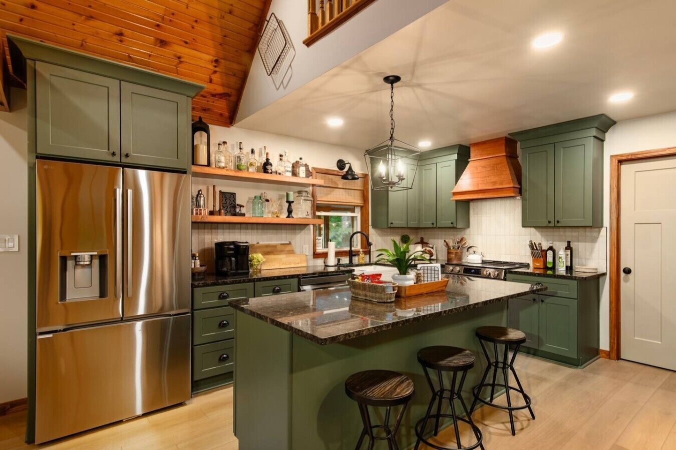 A kitchen with a stainless steel refrigerator, green cabinets, a central island with stools, and a hanging light fixture.