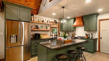 A kitchen with a stainless steel refrigerator, green cabinets, a central island with stools, and a hanging light fixture.