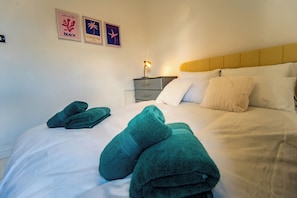 A neatly made bed with white linens and two green towels, a bedside table with a lamp, and wall art depicting beach and Hawaii themes. - 27 Agnes Street (Manchester)