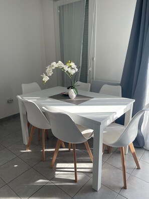 A modern dining area with a white table and chairs, a vase with white flowers, and a view of the outside through a window with a blue curtain. - Nature et ville - Duplex avec terrasse et jardin (Dammarie les lys)