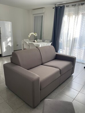 A modern living room with a sofa, a dining table with chairs, and a kitchen area with a refrigerator. - Nature et ville - Duplex avec terrasse et jardin (Dammarie les lys)