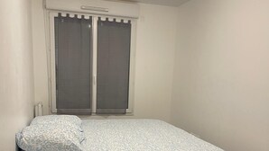 A single bed with a patterned bedspread and pillows, a window with blinds, and a light fixture on the ceiling. - Nature et ville - Duplex avec terrasse et jardin (Dammarie les lys)