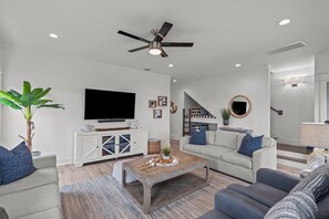 A modern living room with a flat-screen TV, a ceiling fan, and a staircase leading to an upper level. - 1-Min to Beach! New Home! Pets Welcome. 65 Inch Outdoor TV. King Bed! (Galveston)