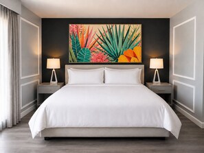 A hotel room with a large bed, two bedside tables with lamps, a colorful tropical plant artwork on the wall, and a view of the outside through a window with curtains.