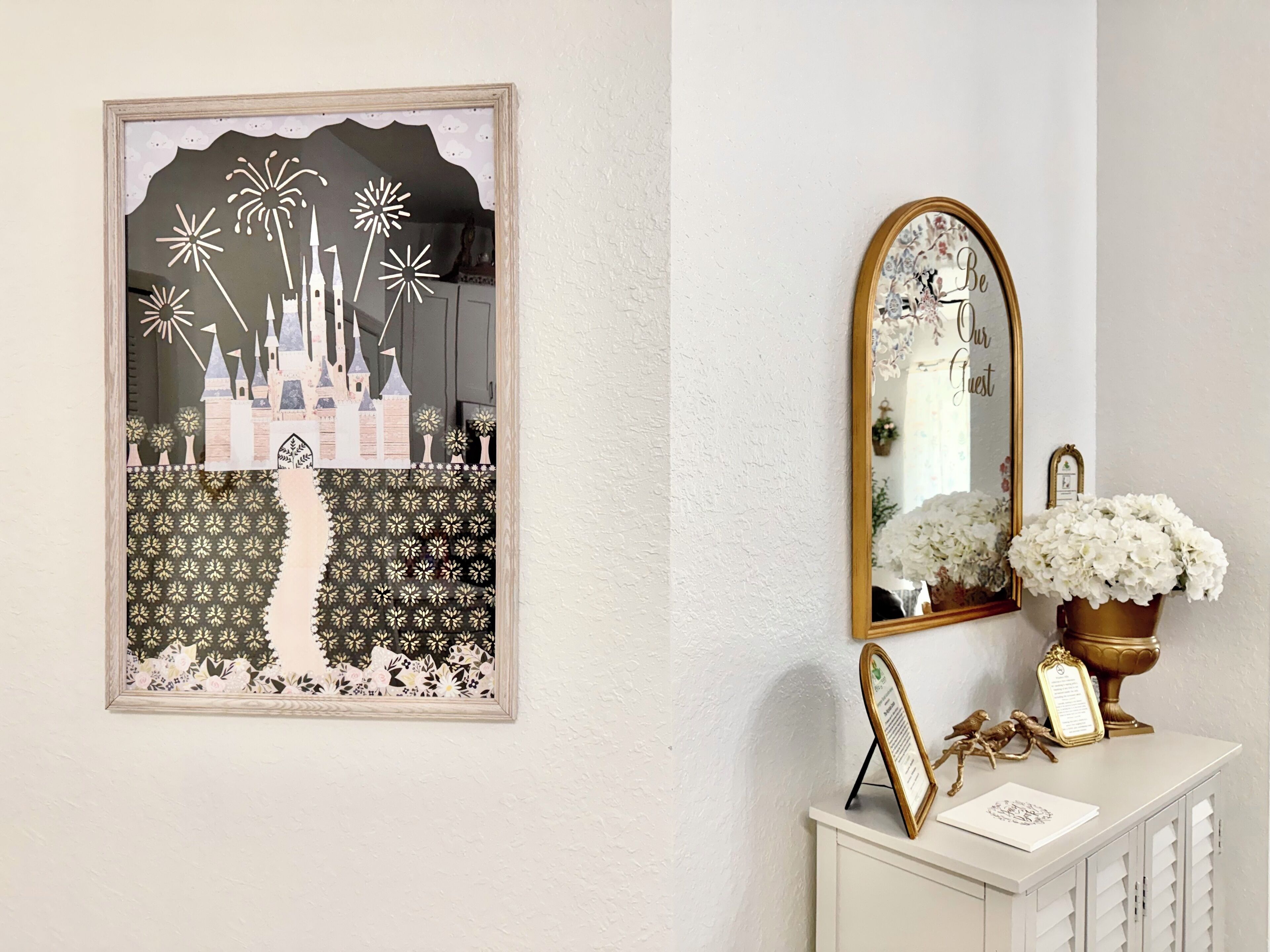 Ein gerahmtes Kunstwerk mit Burg und Löwenzahn, ein Spiegel mit floralem Rahmen und eine weiße Kommode mit goldener Vase und einer gerahmten Speisekarte.