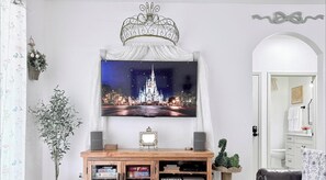 A living room with a flat-screen TV mounted above a wooden console, a decorative metal crown, and a framed picture of a castle at night. - *The Fairytale Condo*  2 Miles to Disney (Kissimmee)