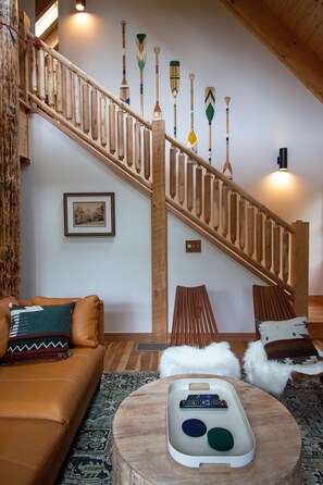 Eine Holztreppe mit Geländer, ein braunes Ledersofa, ein weißer, flauschiger Hocker und eine Fernbedienung auf einem Holztablett.