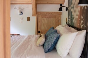 Ein Schlafzimmer mit einem hölzernen Kopfende, einem Bett mit weißer Bettwäsche, einem blau gemusterten Kissen und einer an der Wand montierten Lampe.
