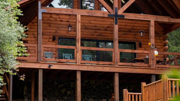 A wooden cabin with a balcony, a stone foundation, and a cross on the roof. - Howdybrook Basecamp | Hocking Hills Cabin (Laurelville)