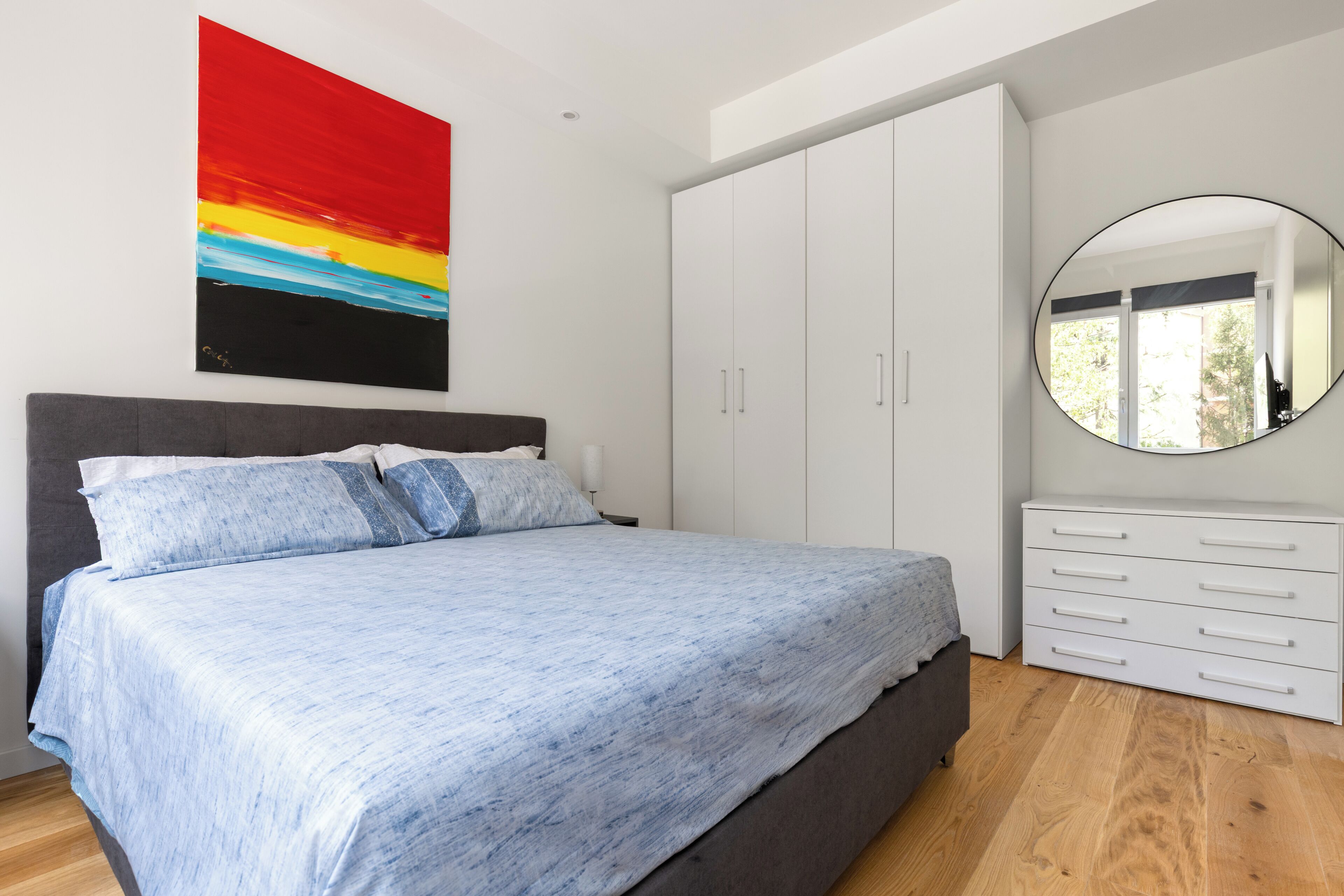Een slaapkamer met een bed, een kledingkast, een spiegel en een schilderij aan de muur.
