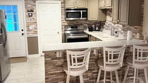 A modern kitchen with a central island, white bar stools, and a stone backsplash.