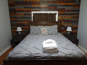 A bed with a grey upholstered headboard, two bedside tables with lamps, a window with curtains, and a wooden wall paneling.