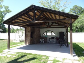 A covered patio area with a brick fireplace, a table with chairs, and a view of a white fence and trees in the background. - House in Savannah, Ga (Garden City)