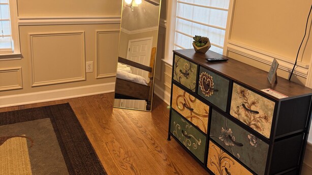 A room with a wooden floor, a mirror, a dresser with drawers, a ceiling fan, and a TV mounted on the wall.