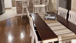 A dining area with a wooden table and white chairs, a kitchen with a stone backsplash, and a white door.