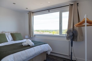 Ein Hotelzimmer mit Bett, Heizkörper, einem Vorhangfenster, einem Standventilator und einem hölzernen Kleiderbügel.