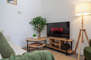 Ein Wohnzimmer mit Fernseher, der 'NETFLIX' anzeigt, einem kleinen runden Tisch mit Fernbedienung, einem grünen Sofa und einer Lampe.