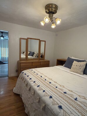 A bedroom with a large bed, a wooden dresser, a ceiling fan, and a window with blinds.