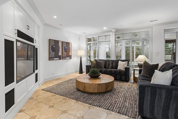 A spacious living room with a large window, a dark grey sofa, a round wooden coffee table, and a patterned rug.