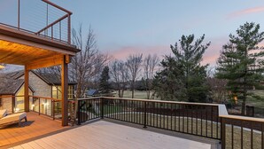 A wooden deck with a railing, overlooking a grassy area with trees and a distant building.