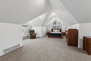 A bedroom with a sloped ceiling, a bed, a chair, a dresser, and a window with blinds.