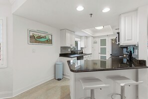 A kitchen with white cabinetry, a black countertop, and a small island with bar stools. - 🧜🏻‍♀️Ocean Front Complex | Ocean Views | Pet Friendly | Las Brisas 523🧜🏻‍♀️ (New Smyrna Beach)