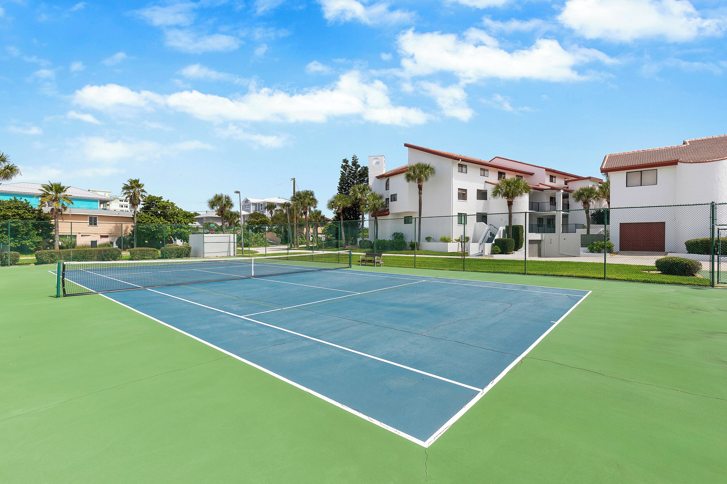 A tennis court with a net, surrounded by a fence and palm trees.