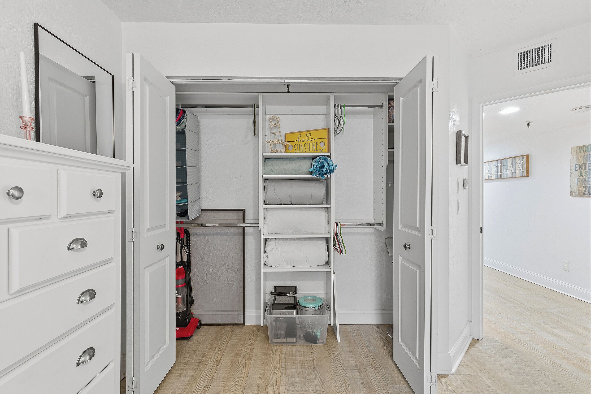 A walk-in closet with open doors, featuring white shelving, a mirror, and a 'hello sunshine' sign.