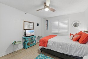 Un dormitorio con una cama, un televisor sobre un mueble, un ventilador de techo y una ventana con persianas.