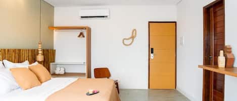 A hotel room with a bed, a wardrobe, a small table, a chair, an air conditioning unit, and a door.