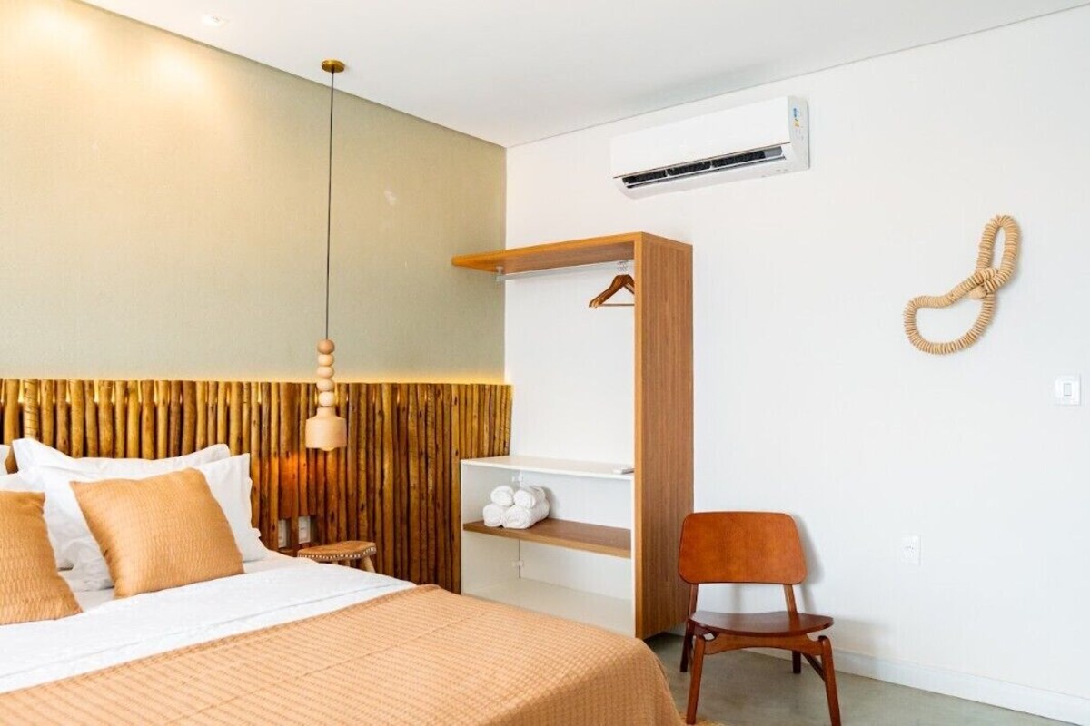 Habitación de hotel con una cama, cabecera de madera, lámpara colgante, una silla de madera, un estante con toallas y un equipo de aire acondicionado.