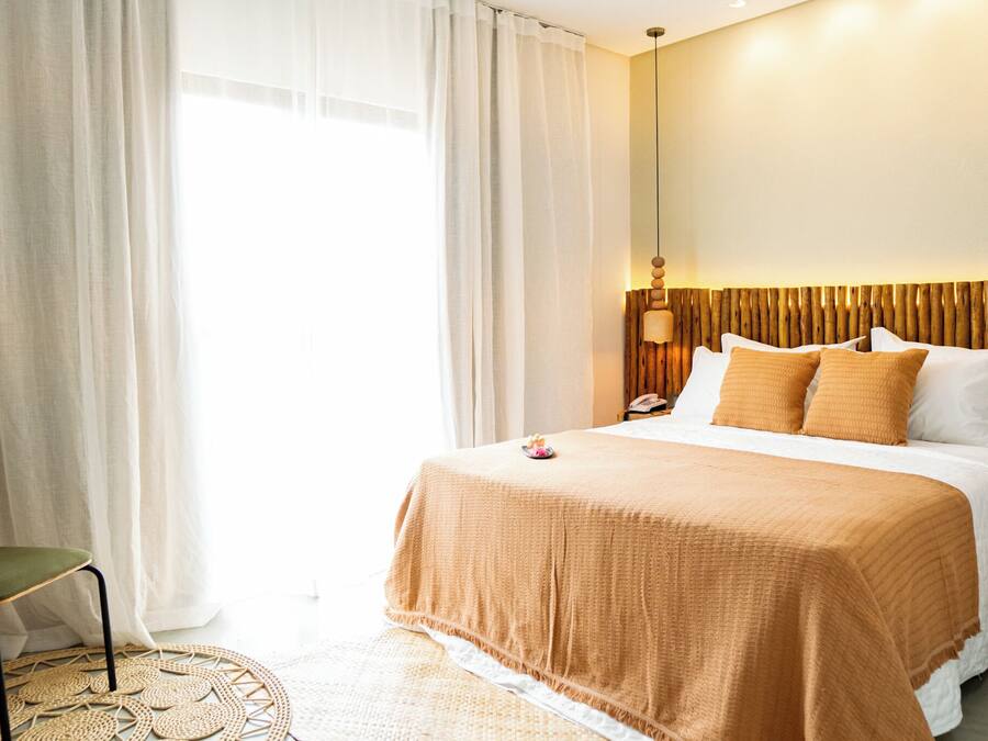 Une chambre d’hôtel avec un lit, une chaise, une table de chevet avec une lampe, et une vue sur l’extérieur à travers des rideaux transparents.