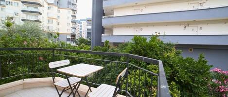 Uma varanda com duas mesas e cadeiras, rodeada por vegetação e com vista para uma área residencial com edifícios altos.
