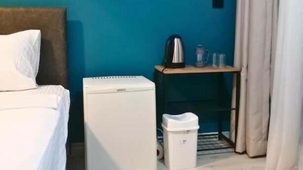 A room with a bed, a small white refrigerator, a trash bin, a shelf with a kettle and cups, and an air conditioning unit on the wall. - Lemon Garden (Antalya)
