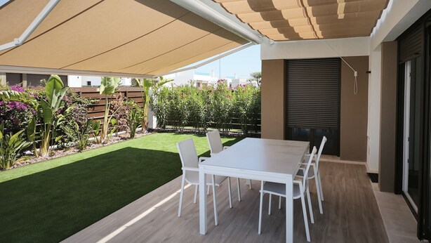 Eine Terrasse im Freien mit Tisch und Stühlen, einem überdachten Bereich und einem Garten mit verschiedenen Pflanzen.