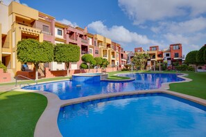 A swimming pool with a surrounding grassy area and a row of multi-story buildings with balconies. - Apartment Royal Park Lomas Cabo Roig (Orihuela)